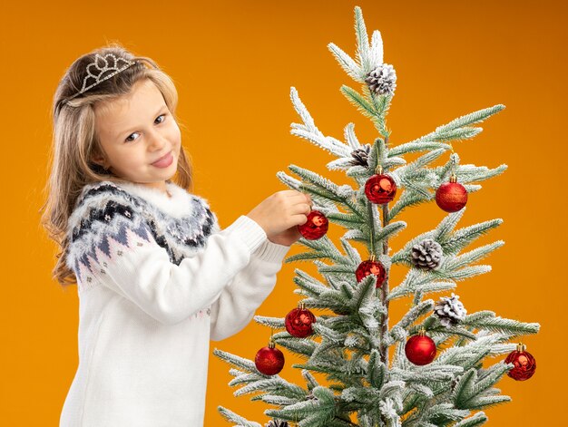 Pleased little girl standing nearby christmas tree wearing tiara with garland on neck hangs toy on the tree isolated on orange background