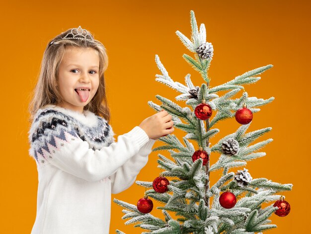 首に花輪とティアラを身に着けているクリスマスツリーの近くに立っている幸せな少女は、オレンジ色の背景で隔離の木におもちゃを掛けます