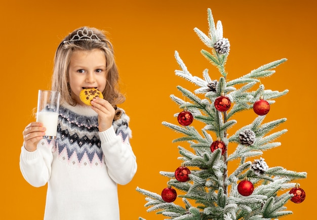 Pleased little girl standing nearby christmas tree wearing tiara with garland on neck holding glass of milk trying cookies isolated on orange background