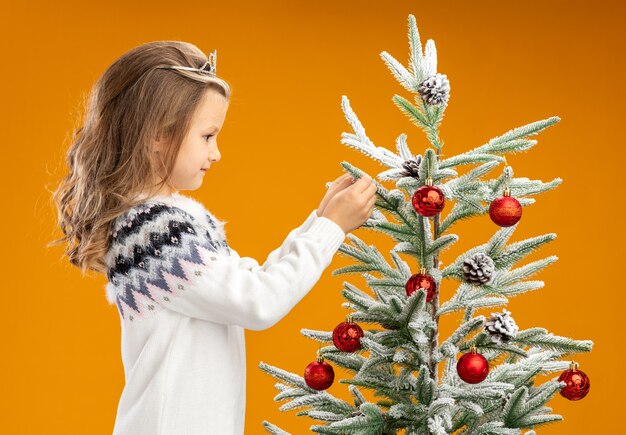 Pleased little girl standing nearby christmas tree wearing tiara with garland on neck holding tree isolated on orange background