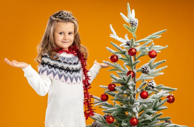 Pleased little girl standing nearby christmas tree wearing tiara with garland on neck spreading hands isolated on orange background