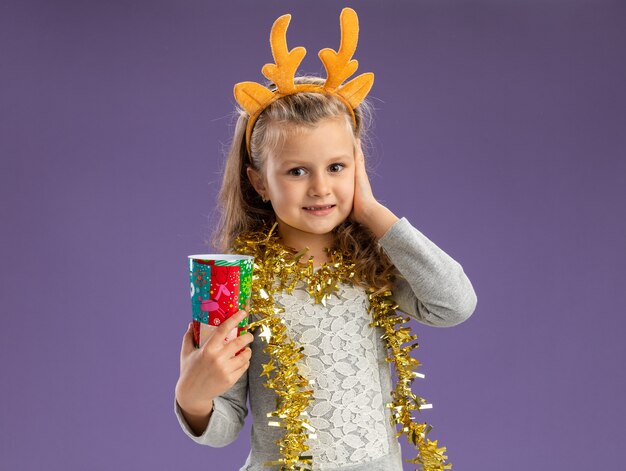 Pleased little girl wearing christmas hair hoop with garland on neck holding christmas cup putting hand on cheek isolated on blue background with copy space