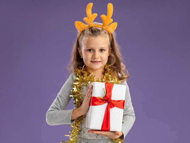 Довольная маленькая девочка в рождественском обруче для волос с гирляндой на шее держит подарочную коробку на синем фоне