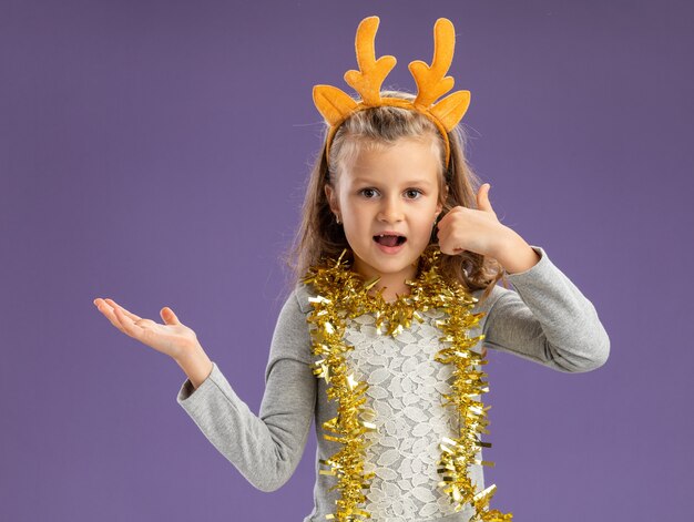 Довольная маленькая девочка в рождественском обруче для волос с гирляндой на шее показывает жест телефонного звонка и указывает на сторону, изолированную на синем фоне с копией пространства