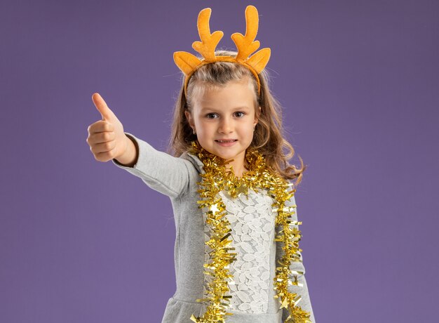 青い背景で隔離の親指を示す首に花輪とクリスマスのヘアフープを身に着けている幸せな少女