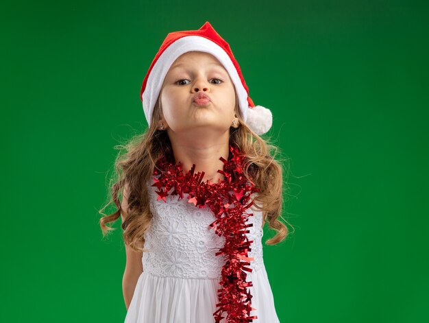 Pleased little girl wearing christmas hat with garland on neck showing kiss gesture isolated on green background