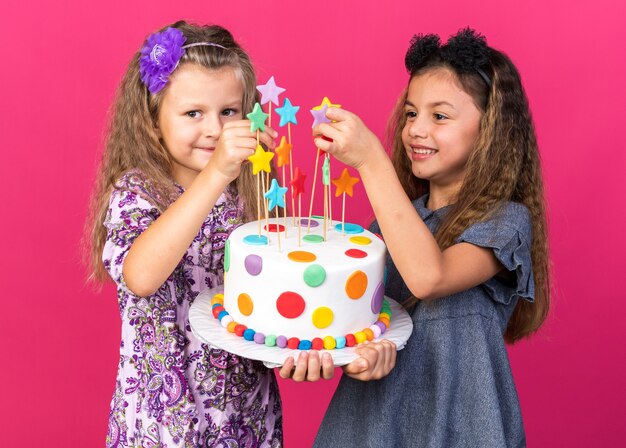 コピースペースとピンクの壁に分離されたバースデーケーキを一緒に保持しているかわいい女の子を喜ばせ