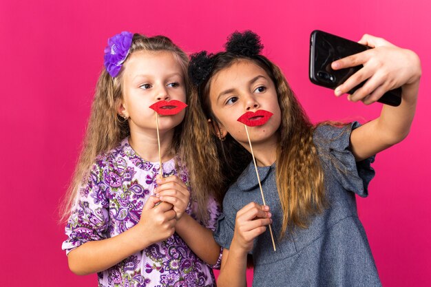 Довольные маленькие красивые девушки держат поддельные губы на палках, делающие селфи на розовой стене с копией пространства