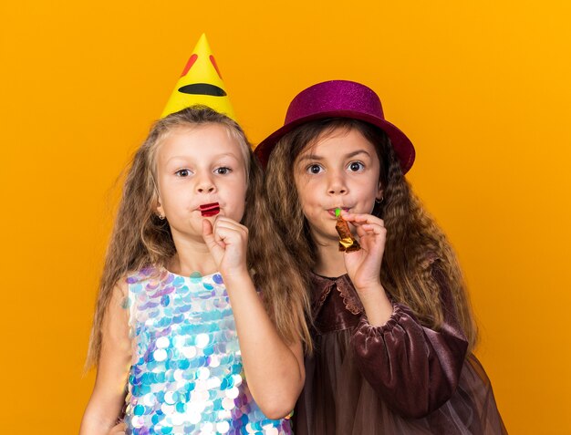 pleased little pretty girls with party hats blowing party whistles isolated on orange wall with copy space