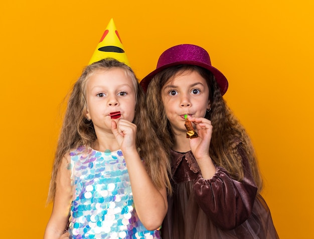 Довольные маленькие симпатичные девушки в шляпах для вечеринок дуют в свистки на оранжевой стене с копией пространства