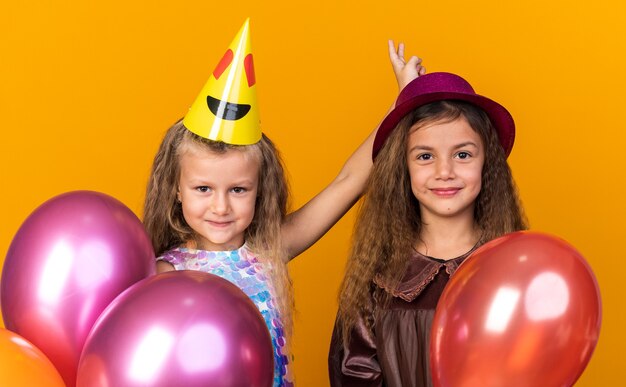 Довольные маленькие симпатичные девушки в праздничных шляпах с гелиевыми шарами, изолированными на оранжевой стене с копией пространства