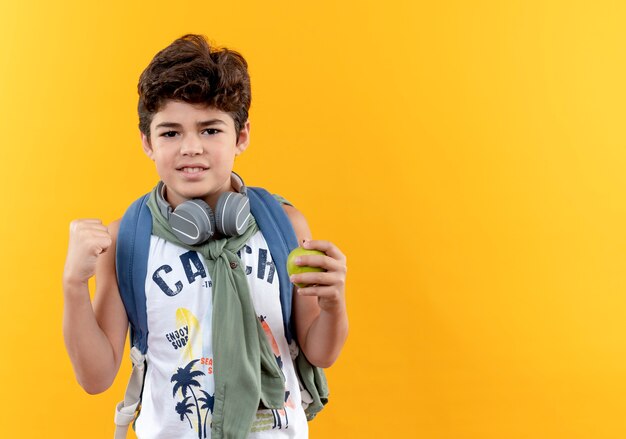 Pleased little school boy wearing back bag and headphones holding apple and showing yes gesture isolated on yellow background with copy space