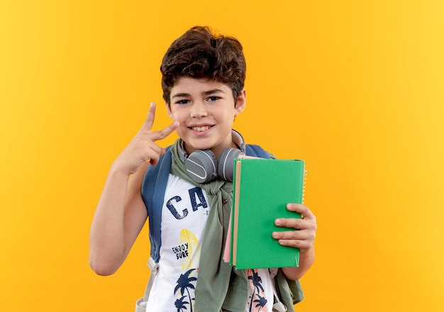 本を保持し、黄色の背景に分離された平和のジェスチャーを示すバックバッグとヘッドフォンを身に着けている小さな男子生徒を喜ばせます