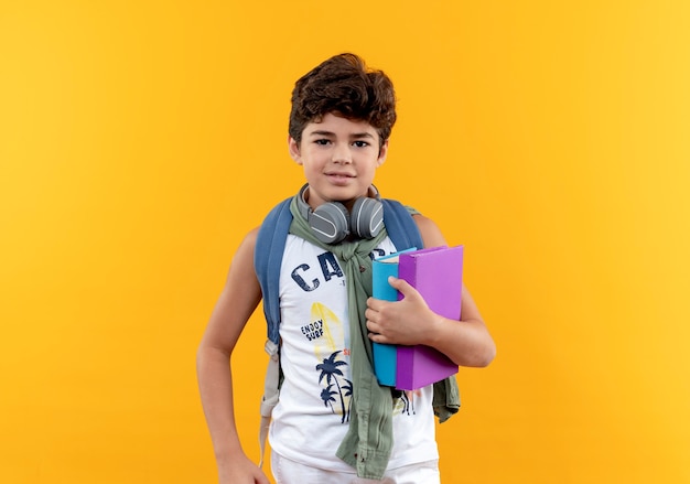 Довольный маленький школьник в задней сумке и наушниках держит книги и кладет руку на бедро, изолированные на желтом фоне