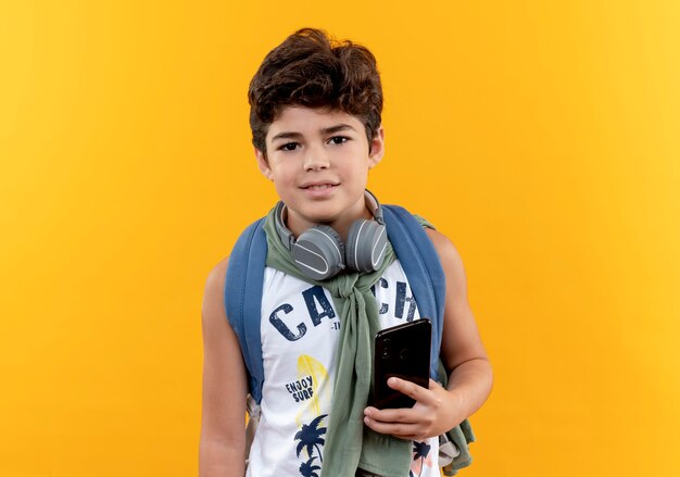 Pleased little school boy wearing back bag and headphones holding phone isolated on yellow background
