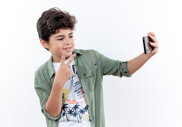 Pleased little schoolboy showing peace gesture and take selfie isolated on white background