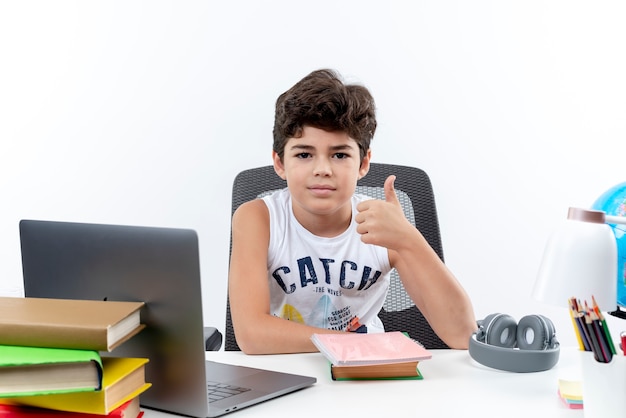 Pleased little schoolboy sitting at desk with school tools his thumb up isolated on white background