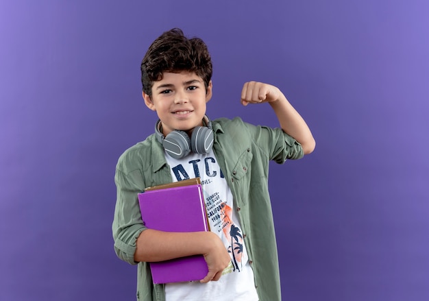 Довольный маленький школьник в наушниках держит книги и делает сильный жест, изолированный на фиолетовой стене