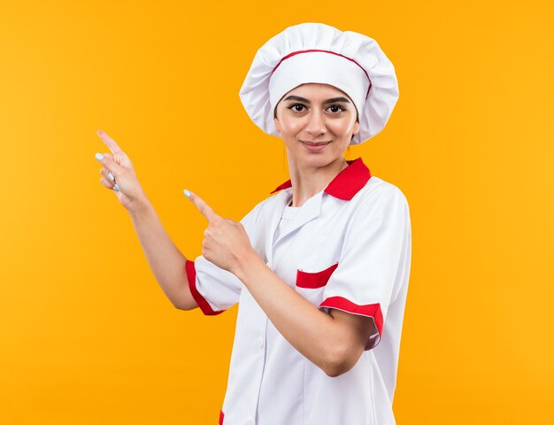 Pleased looking at camera young beautiful girl in chef uniform points at side 