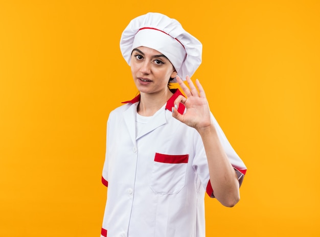 Piacere guardando la telecamera giovane bella ragazza in uniforme da chef che mostra un gesto ok