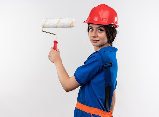 Довольно глядя на камеру молодая женщина-строитель в униформе, держащая роликовую щетку