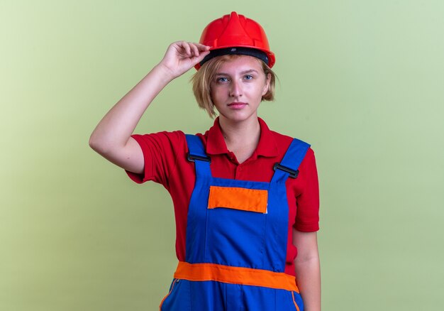 pleased looking at front young builder woman in uniform isolated on olive green wall