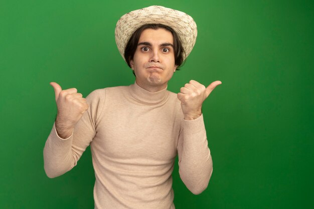 Free photo pleased looking at front young handsome guy wearing hat showing thumbs up isolated on green wall with copy space