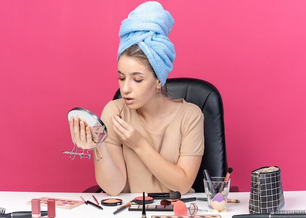 Pleased looking at mirror young beautiful girl sits at table with makeup tools wrapped hair in towel applying lipgloss isolated on pink wall