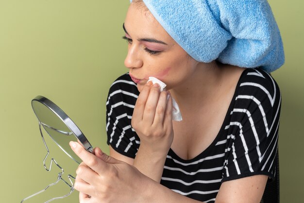 Free photo pleased looking at mirror young beautiful girl sits at table with makeup tools wrapped hair in towel wiping face with napkin isolated on olive green wall