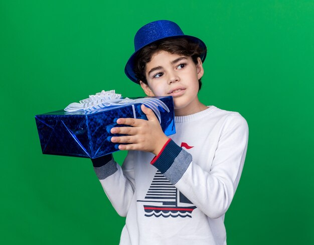 Pleased looking side little boy wearing blue party hat holding gift box 