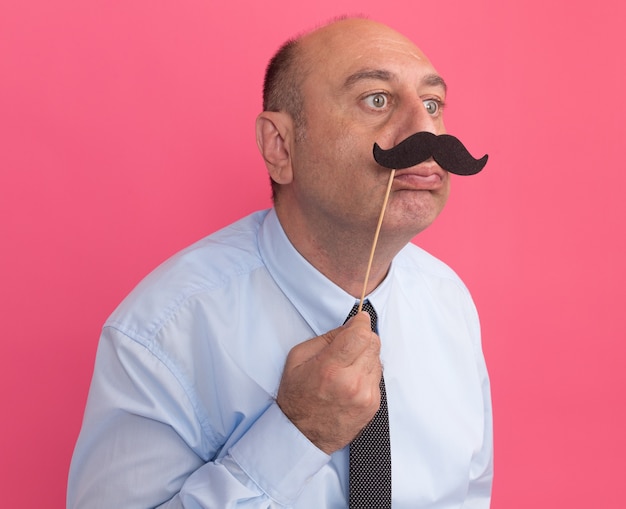 Foto gratuita soddisfatto guardando un uomo di mezza età laterale che indossa una maglietta bianca con cravatta che tiene baffi finti sul bastone isolato sul muro rosa