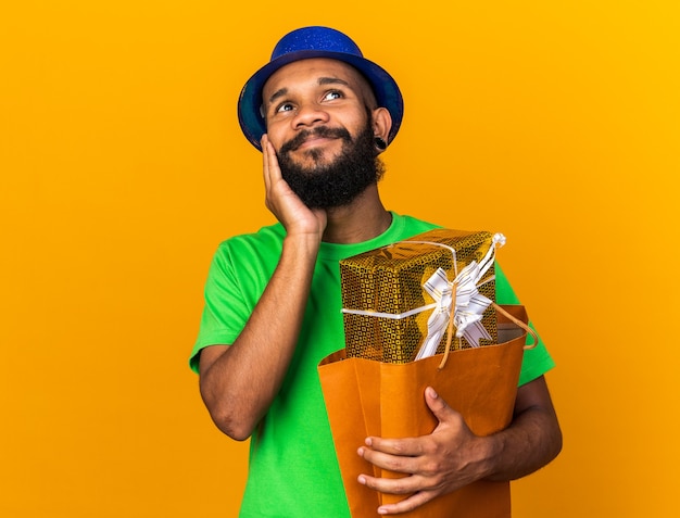주황색 벽에 격리된 손으로 뺨을 덮은 선물 가방을 들고 파티 모자를 쓴 젊은 아프리카계 미국인 남자