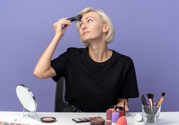 Pleased looking side young beautiful girl sits at table with makeup tools applying powder blush with powder brush isolated on blue wall