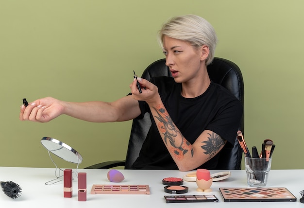 Piacevole lato guardando la giovane bella ragazza si siede al tavolo con gli strumenti per il trucco tenendo fuori l'eyeliner a lato isolato sul muro verde oliva