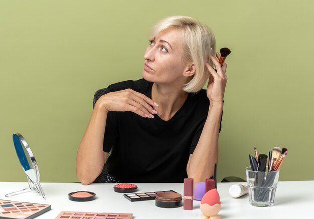 Pleased looking side young beautiful girl sits at table with makeup tools holding powder brush isolated on olive green wall