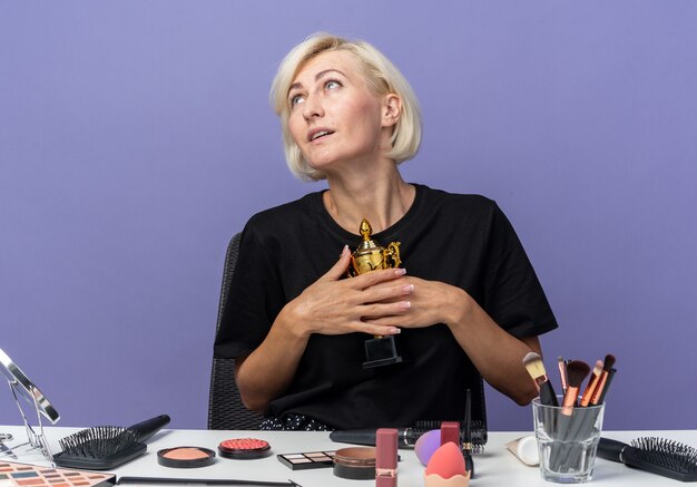 Pleased looking side young beautiful girl sits at table with makeup tools holding winner cup isolated on blue wall