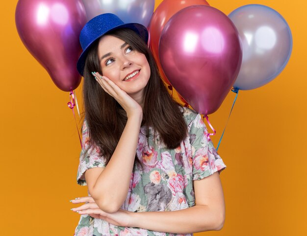 Pleased looking side young beautiful girl wearing party hat standing in front balloons putting hand on cheek isolated on orange wall