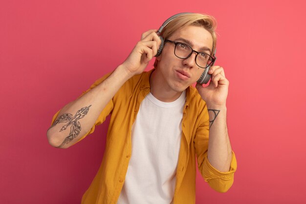 Pleased looking at side young blonde guy wearing yellow t-shirt and glasses with headphones isolated on pink