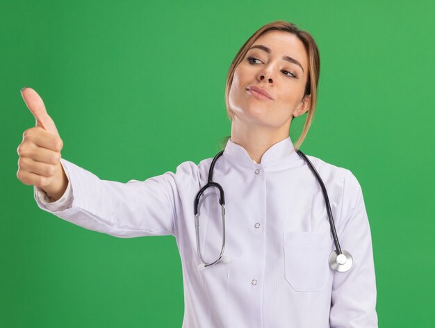 녹색 벽에 고립 된 엄지 손가락을 보여주는 청진 기 의료 가운을 입고 측면 젊은 여성 의사를보고 기쁘게 생각