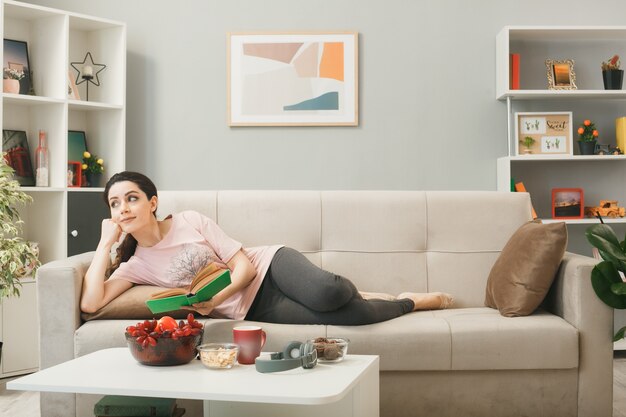 Pleased looking side young girl lying on sofa behind coffee table holding book in living room