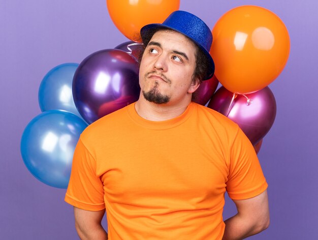 Pleased looking side young man wearing party hat standing in front balloons isolated on purple wall