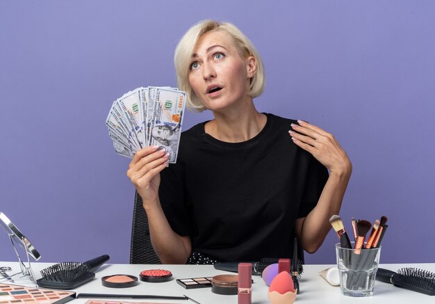 青い壁に分離された現金を保持している化粧ツールでテーブルに座っている若い美しい少女を見て喜んで