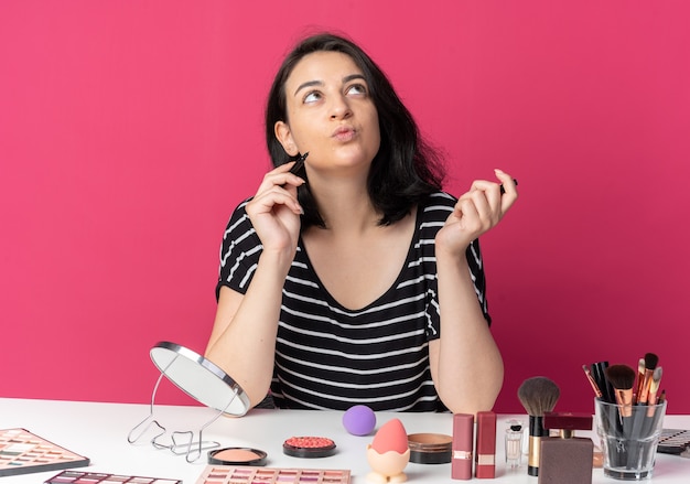 Pleased looking up young beautiful girl sits at table with makeup tools holding eyeliner isolated on pink wall