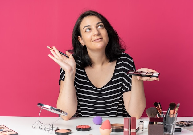 Pleased looking up young beautiful girl sits at table with makeup tools holding eyeshadow palette with makeup brush isolated on pink wall