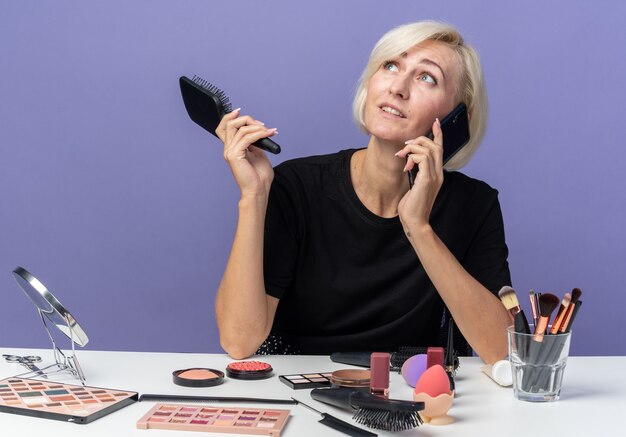 Pleased looking up young beautiful girl sits at table with makeup tools speaks on phone holding comb isolated on blue wall