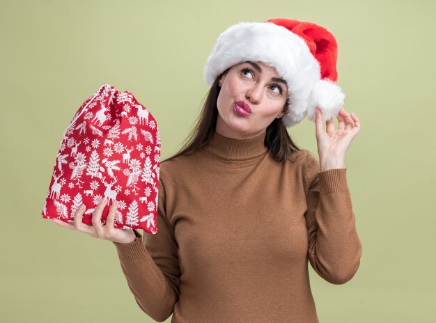 オリーブグリーンの背景に分離されたクリスマスバッグを保持しているクリスマス帽子をかぶって若い美しい少女を探して喜んで