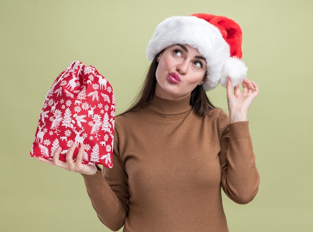 オリーブグリーンの背景に分離されたクリスマスバッグを保持しているクリスマス帽子をかぶって若い美しい少女を探して喜んで