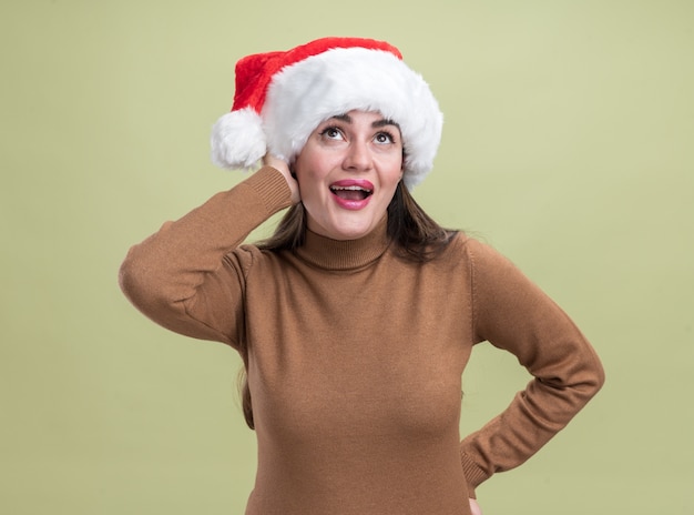 Pleased looking at up young beautiful girl wearing christmas hat putting hands on cheek and hip isolated on olive green background