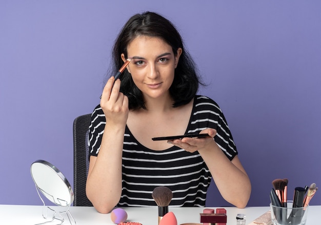Pleased looking  young beautiful girl sits at table with makeup tools applying eyeshadow with makeup brush isolated on blue wall