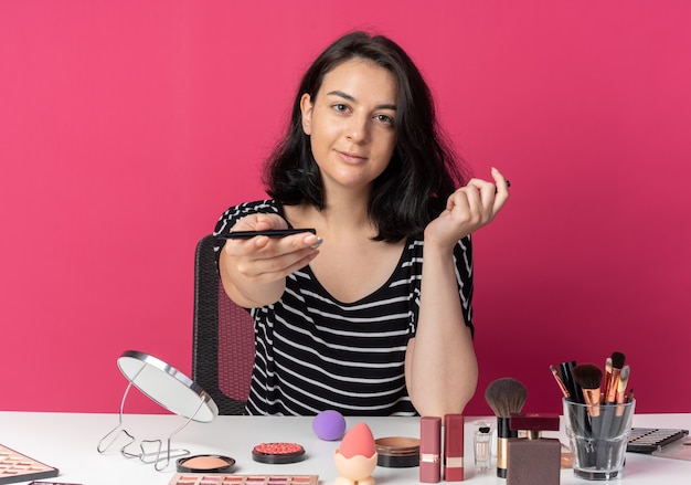 Pleased looking  young beautiful girl sits at table with makeup tools holding out eyeliner  isolated on pink wall